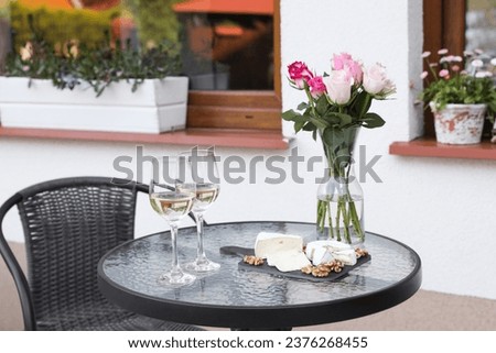 Vase with roses, glasses of wine and food on glass table near house on outdoor terrace