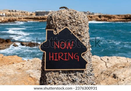 Now hiring symbol. Concept words Now hiring on beautiful black chalk blackboard. Beautiful stone sea blue sky background. Business marketing, motivational now hiring concept. Copy space.