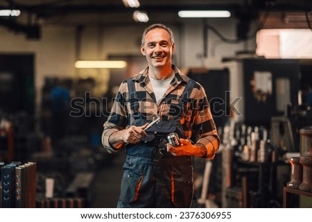 A picture of a metallurgy mechanic holding a tool and a cylinder machine element, smiling at the camera. Blurred background of manufacture facility with various metal shapes and machines. Royalty-Free Stock Photo #2376306955