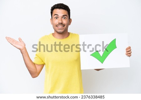 Young handsome man over isolated white background holding a placard with text Green check mark icon with surprised expression