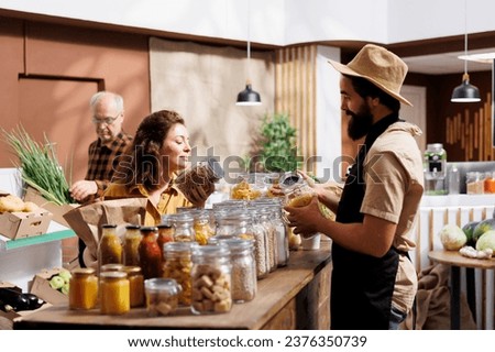 Woman shopping in zero waste grocery store, looking at bulk products in reusable packaging while being assisted by helpful clerk. Customer in local neighborhood picking ethically produced food Royalty-Free Stock Photo #2376350739