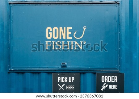 A blue metal wall window with gone fishing painted in yellow text and a fishing hook symbol.  Below the door there are two black signs with pick up here and order here. There's a knife and fork symbol