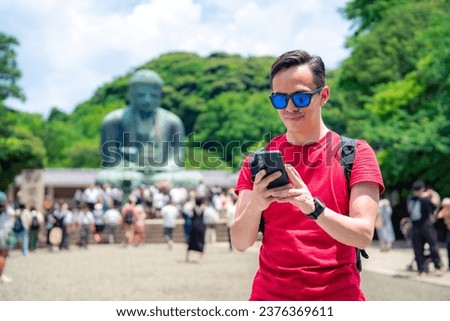 Full body side view of Hispanic young male in casual clothes with backpack while taking picture with smartphone of Kotoku-in Buddhist temple structure in Kamakura Tokyo, Japan