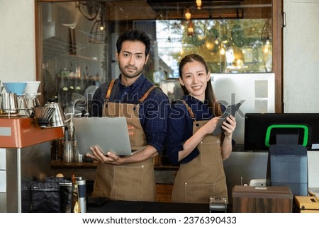 Asian couple or indian in aprons looking camera and checking online orders together, husband and wife open and share small business cofee shop. Owners using laptops in their restaurants talk customer. Royalty-Free Stock Photo #2376390433