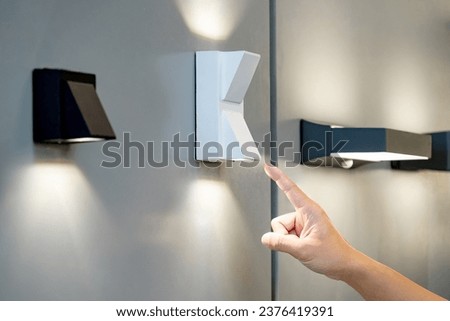 Designer hand pointing at sample of exterior LED wall lamp in home design store. Modern white uplight and downlight light fixture mounted on wall for outdoor lighting Royalty-Free Stock Photo #2376419391
