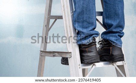 Male maintenance worker feet with protective safety shoes climbing up on aluminum step ladder at construction site. Building service tool and equipment Royalty-Free Stock Photo #2376419405
