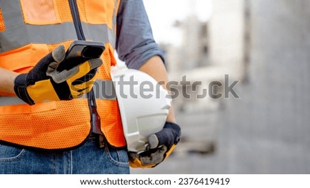 Construction worker man with orange reflective vest holding white protective safety helmet using smartphone at unfinished building site. Male engineer or foreman stay connected with social media app