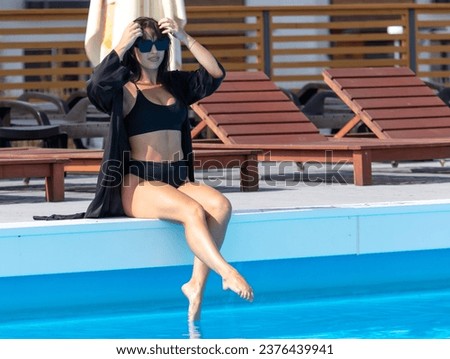 A girl in a black swimsuit and glasses sits by the pool.