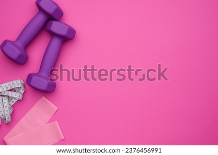 Layout of two rubberized dumbbells, measuring tape, fitness elastic band on a pink background, top view