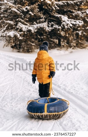 Active toddler boy in a yellow jacket pulls a rope snow tubing uphill in snow-covered forest.Winter fun,active lifestyle concept.