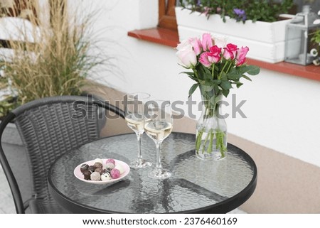Vase with roses, glasses of wine and candies on glass table near house on outdoor terrace