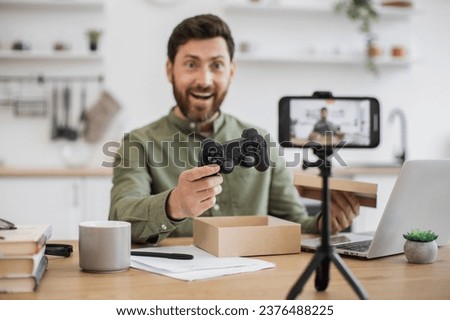 Thrilled male vlogger looking at mobile camera with wireless joystick in hand after opening cardboard box. Bearded man sharing sincere emotions with followers of parcel unpacking during live stream. Royalty-Free Stock Photo #2376488225