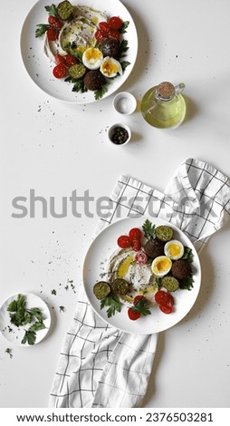 hummus with falafel, boiled egg, cherry tomatoes and parsley on plate, ingredients on table, appetizing vegetarian healthy dish, white background, top view, flat lay Royalty-Free Stock Photo #2376503281