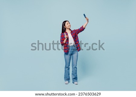 Full body young woman of Asian ethnicity 20s wear checkered shirt doing selfie shot on mobile cell phone post photo on social network show v-sign isolated on plain pastel light blue color background.