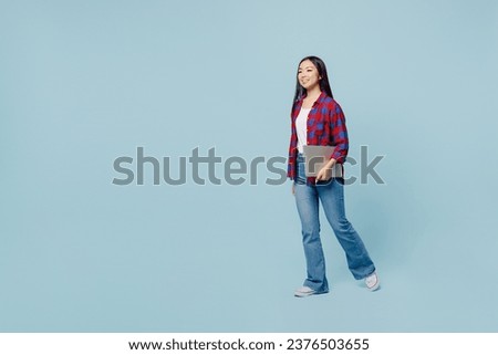 Full body young smiling fun woman of Asian ethnicity wear checkered shirt hold closed laptop pc computer walking going isolated on plain pastel light blue color background. People lifestyle concept