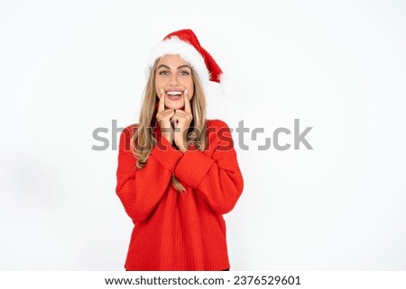 Happy Beautiful hispanic woman wearing christmas hat and red knitted sweater with toothy smile, keeps index fingers near mouth, fingers pointing and forcing cheerful smile