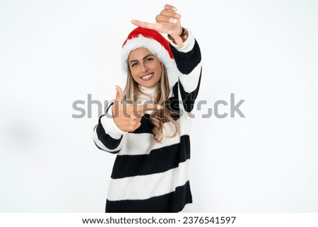Beautiful hispanic woman wearing christmas hat and striped knitted sweater making finger frame with hands. Creativity and photography concept.