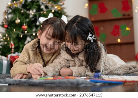 A cute and happy young Asian girl is helping her younger sister making a Christmas card or painting a picture with pencil colors while lying on the living room's floor together. Merry Christmas