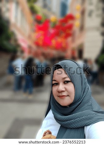 Muslim woman wearing a hijab, taking a selfie outdoors with a blurred background.