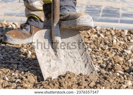Digging gravel with a wide shovel on a construction site,The worker rests his foot on the shovel to push it into the gravel and scoop the material onto it. Royalty-Free Stock Photo #2376557147