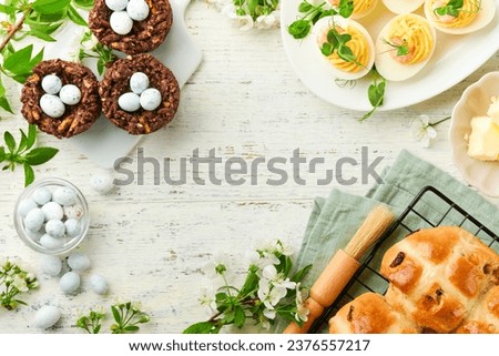 Easter brunch or breakfast. Easter chocolate nest cake with chocolate candy eggs, traditional hot cross buns and deviled eggs with bouquets of blooming apple trees. Spring Easter holiday food concept. Royalty-Free Stock Photo #2376557217