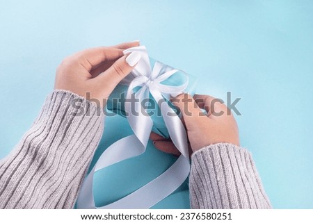 First person top view photo of female hands holding blue gift box with white ribbon. A pastel blue setting welcomes your words. my hands cradle a pastel blue gift box