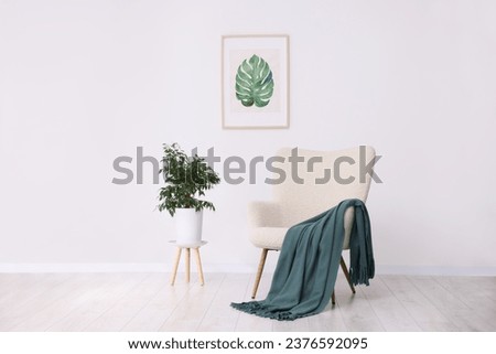 Comfortable armchair, blanket, houseplant and picture on white wall indoors Royalty-Free Stock Photo #2376592095