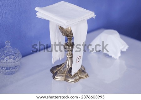 golden metal chalice or goblet with symbol of the cross of Christ, covered with white embroidered fabric on the altar - ampulla Royalty-Free Stock Photo #2376603995