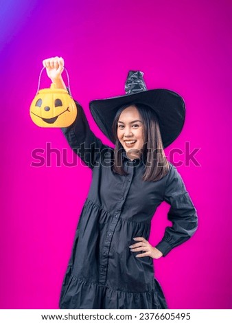 Portrait of an Asian Indonesian woman wearing a Halloween-themed costume with a witch hat, smiling while holding a pumpkin. Isolated against a magenta background.