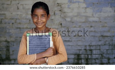 Portrait of happy cute little indian girl in school uniform holding blank slate, Adorable elementary kid showing black board. child education concept. rural india. Royalty-Free Stock Photo #2376610285