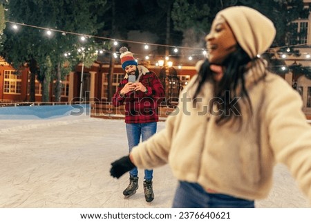 A young girl is ice skating in the skating ring and her boyfriend is taking a picture of her. They are having fun on a winter evening.