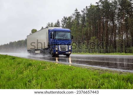 A truck with a semi-trailer transports cargo in summer in rainy weather against the background of a forest. Import and export of goods. Trucking business. Copy space for text Royalty-Free Stock Photo #2376657443
