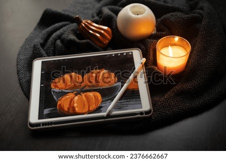 The still life picture with pumpkins drawn on the electronic tablet next to burning candle and ceramic pumpkin figurine on black table. Inspiration, creativity, hobby. Halloween, Thanksgiving day