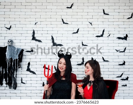 Speak of the devil with Asian evil lady holding red trident and witch girl look at a prop and talk about someone in Horror Halloween theme decorate vampire bat paper cut and creepy ghost on brick wall Royalty-Free Stock Photo #2376665623