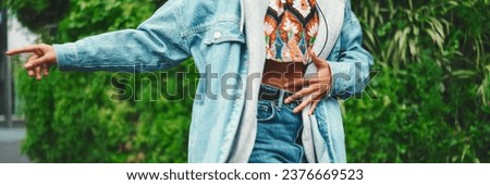Closeup, smiling girl wearing denim jacket, in crop top with national pattern walks in city park listening to music on headphones and dancing on green trees background.