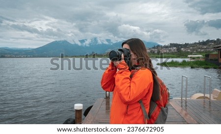 Beautiful young Latin American woman dressed in orange jacket with backpack seen from the front taking photos on a wooden pier on a lake surrounded by mountains on a cloudy day