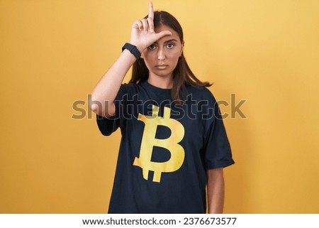 Young hispanic woman wearing bitcoin t shirt making fun of people with fingers on forehead doing loser gesture mocking and insulting. 