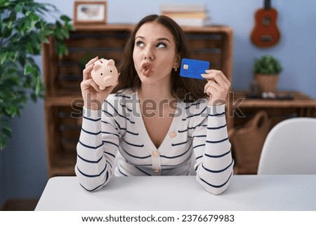 Young hispanic girl holding piggy bank and credit card making fish face with mouth and squinting eyes, crazy and comical. 