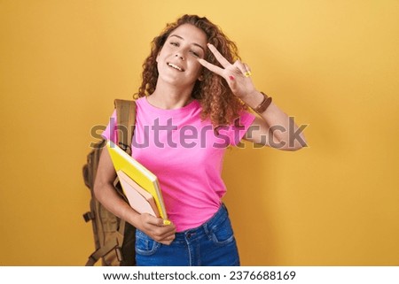 Young caucasian woman wearing student backpack and holding books doing peace symbol with fingers over face, smiling cheerful showing victory 