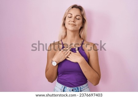 Young blonde woman standing over pink background smiling with hands on chest with closed eyes and grateful gesture on face. health concept. 
