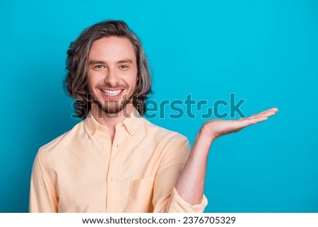 Portrait of toothy beaming guy with long haircut wear stylish shirt palm presenting object empty space isolated on blue color background