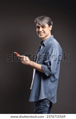 Portrait of Brazilian man, wearing button-up shirt and jeans, profile position looking at camera and holding smartphone, gray hair - Color photo - Brazil