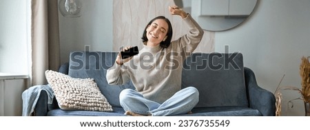 Happy and carefree girl singing and listening to music on smartphone app, using mobile phone as microphone, sitting on sofa. Royalty-Free Stock Photo #2376735549