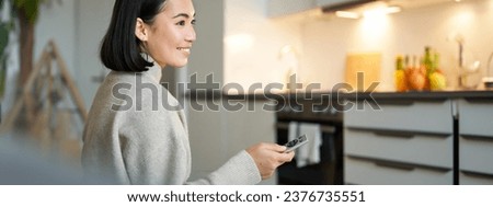 Close up portrait of smiling asian woman watching television, sitting on sofa with remote and switching chanel, looking relaxed.