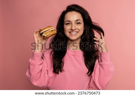 Waist-up portrait of a cheerful brunette with a double cheeseburger in her hand looking at the camera during the studio photo shoot. Fast food consumption concept
