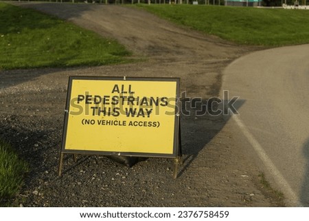 Vibrant yellow directional sign placed beside a gravel and paved split pathway, reading 'all pedestrians this way (no vehicle access)'.
