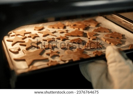 Hand in oven mitts putting tray with gingerbread cookies in christmas festive shapes in modern oven in white kitchen. Making christmas gingerbread cookies Royalty-Free Stock Photo #2376764675