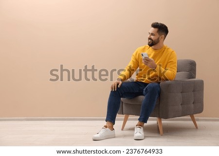 Handsome man with smartphone sitting in armchair near beige wall indoors, space for text Royalty-Free Stock Photo #2376764933
