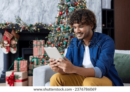 A man uses a tablet computer while sitting at home in the living room for Christmas. A Latin American uses the app, watches a video, smiles, celebrates New Year's holidays near the Christmas tree.