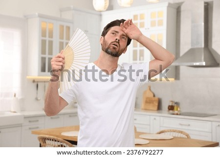 Bearded man waving white hand fan to cool himself in kitchen